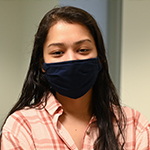 Brown female (Meghan Gupta) with long-length brown hair wearing a light plaid shirt and a black mask covering her nose and mouth